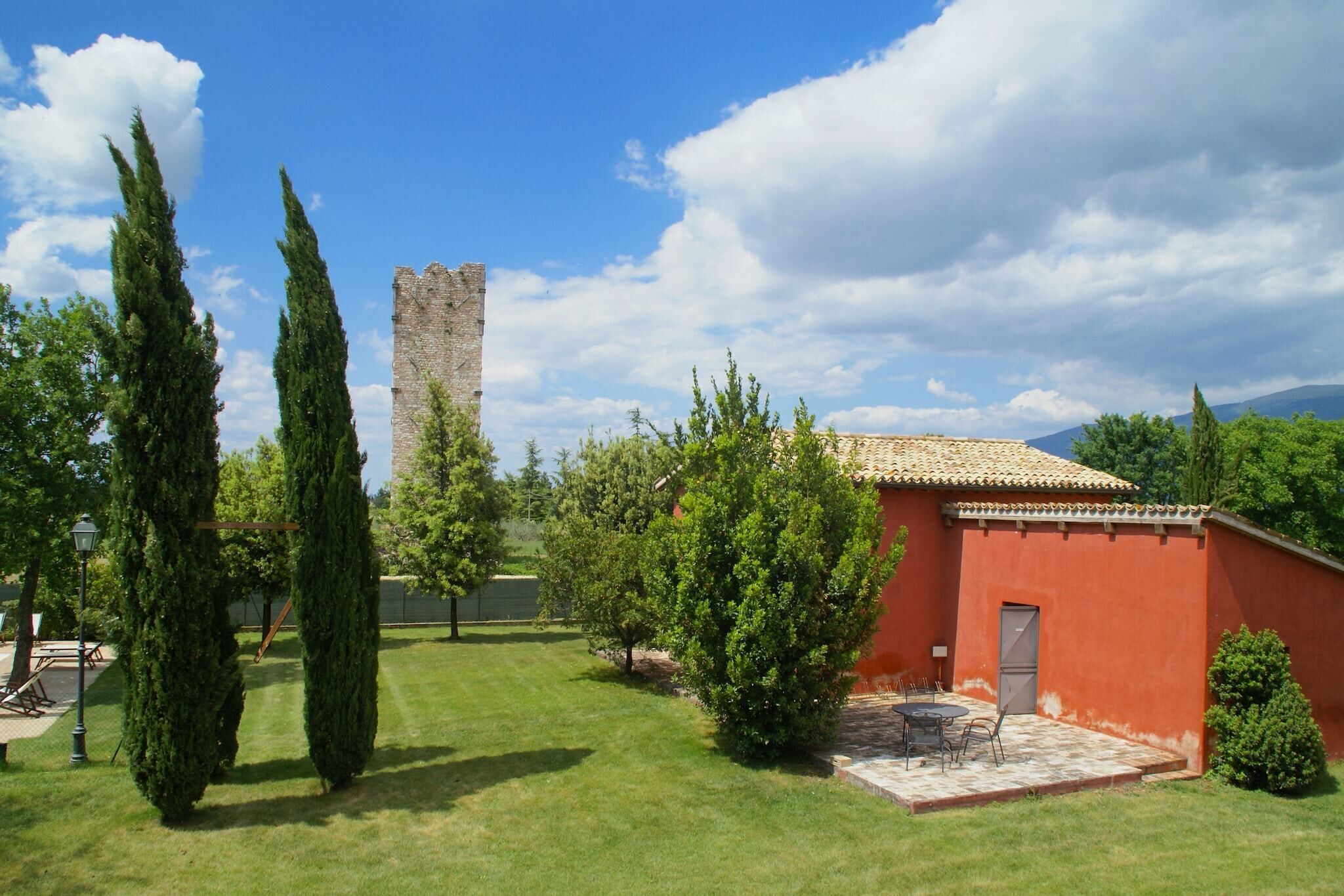 A courtyard with a red building, a tall tower, and cypress trees.