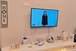 Une pièce avec un téléviseur fixé au mur qui diffuse une image d’une personne, une étagère blanche contenant divers objets et un panneau mural avec le mot « MAISON ».