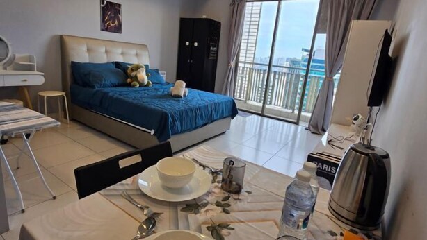 Habitación de hotel con cama, escritorio, silla, mesa con un tazón y una botella, y vista a la ciudad a través de la ventana.