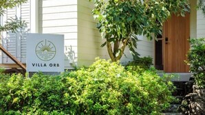 Une enseigne indiquant « Villa Orb », avec un logo, entourée d’une végétation luxuriante et d’une porte en bois.