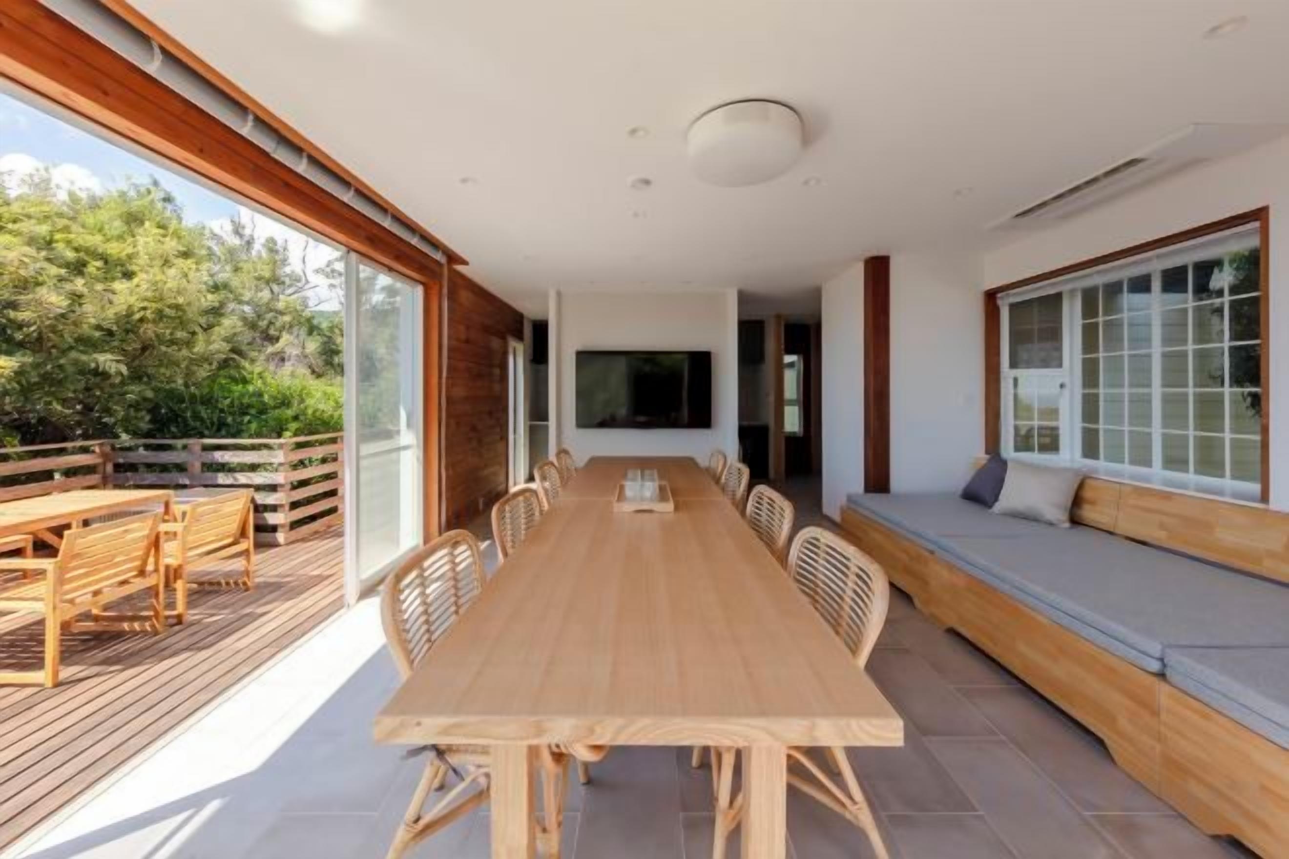 A spacious dining area with a long wooden table and chairs, a built-in bench along the wall, and large windows providing natural light.