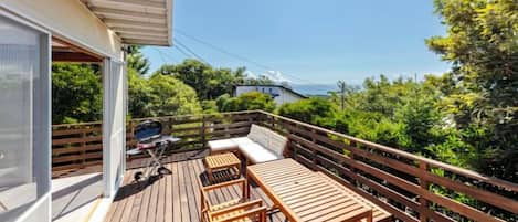 Una terraza de madera con mobiliario exterior, una parrilla y vistas al entorno verde.
