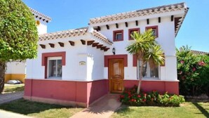 A two-story building with a red and white facade, a wooden door, and a small garden with a palm tree and flowers. - Villa Dionysus- 2 Bedroom, 2 Bathroom Villa, south facing with private pool (Torre-Pacheco)