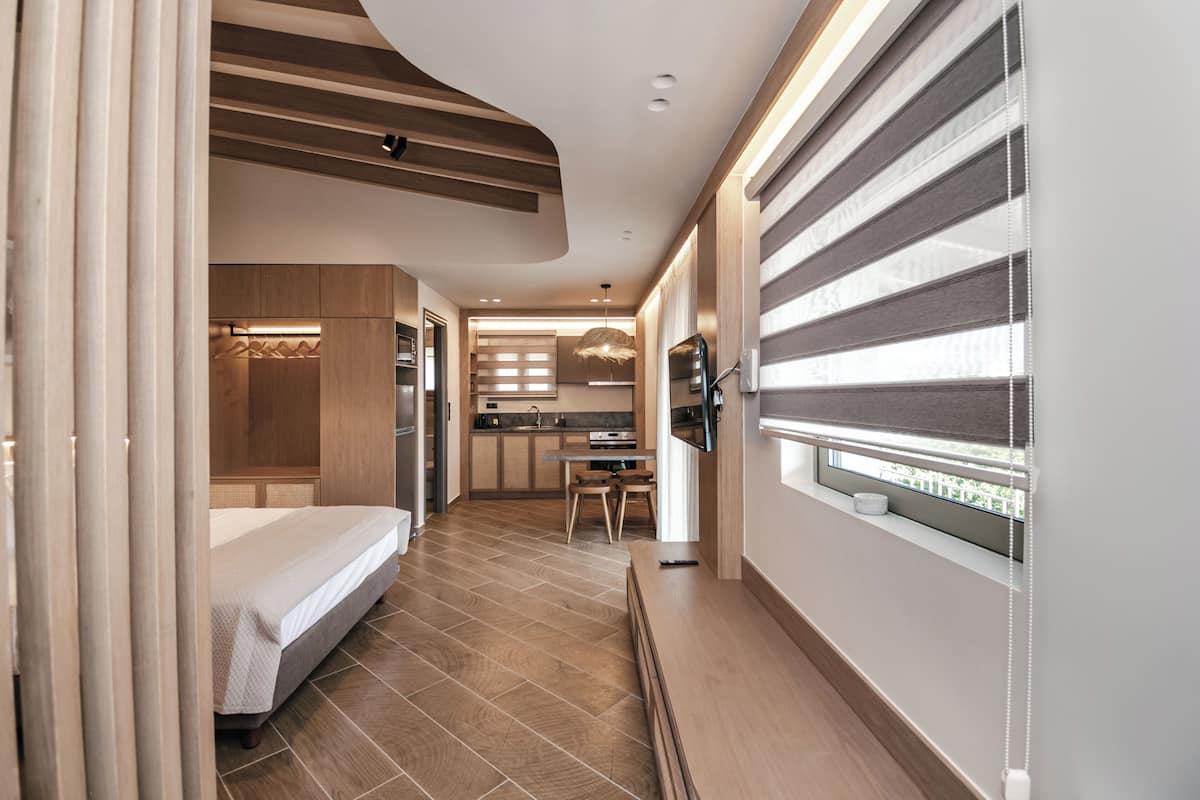A modern hotel room with a bed, a small kitchenette, a dining area, and a window with blinds.
