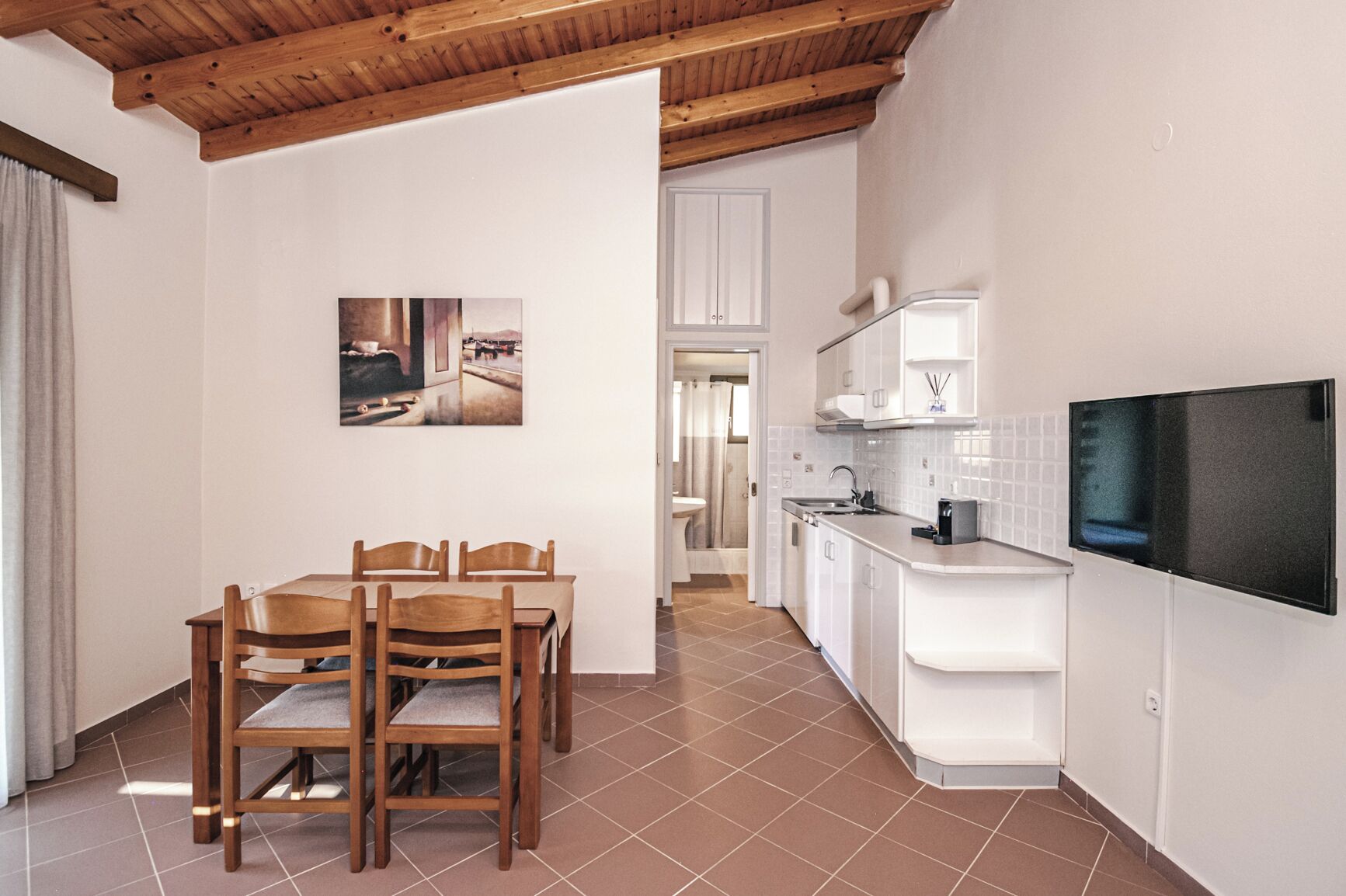 A modern kitchen with white cabinets, a dining area with wooden chairs, and a flat-screen TV mounted on the wall.