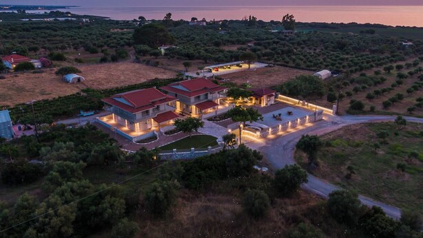 A large, multi-story house with a red roof, surrounded by greenery and a view of the sea at dusk. - Terra Hélios Suites & Studios (Trifylia)