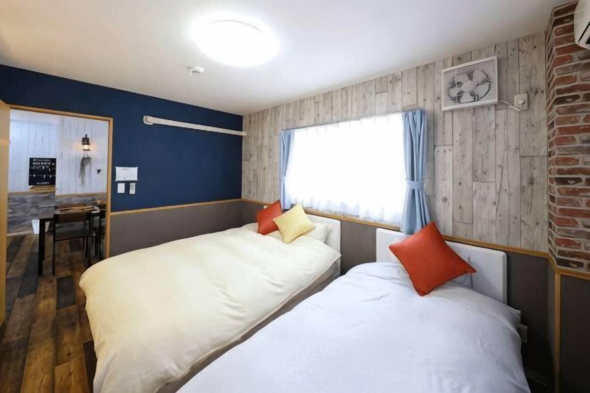 A room with two beds, a wooden wall, a window with curtains, a clock, and a small table with chairs.