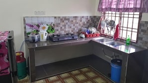 A kitchen with a tiled backsplash, a gas stove, a sink, and a window with a curtain. - Homestay Che Wan (Kuala Kuantan)