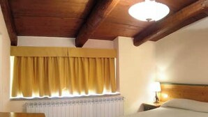 A hotel room with a wooden ceiling, a bed with white bedding, a nightstand with a lamp, and a window with yellow curtains. - Apartment 26 with 1 Bedroom and Bathroom - Simple and Comfortable (Castel del Piano)