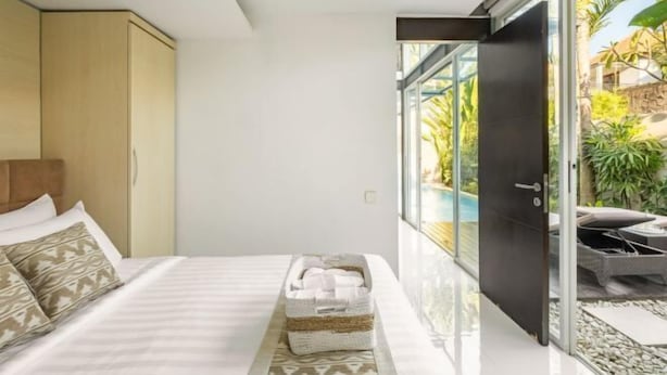 A modern hotel room with a bed, a small bedside table, a glass door leading to a balcony with outdoor furniture. - Villa De Luca By Aishwarya Bali International (Bali)