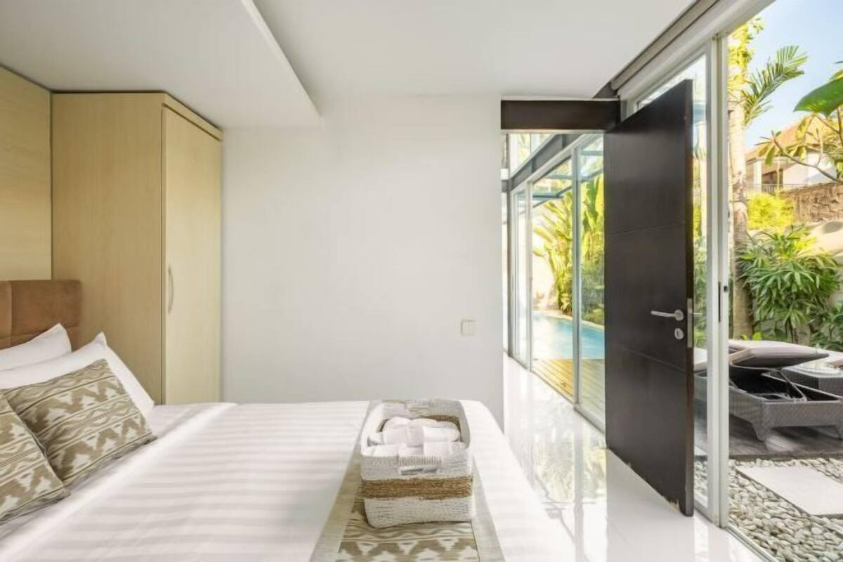 A modern hotel room with a bed, a small bedside table, a glass door leading to a balcony with outdoor furniture.