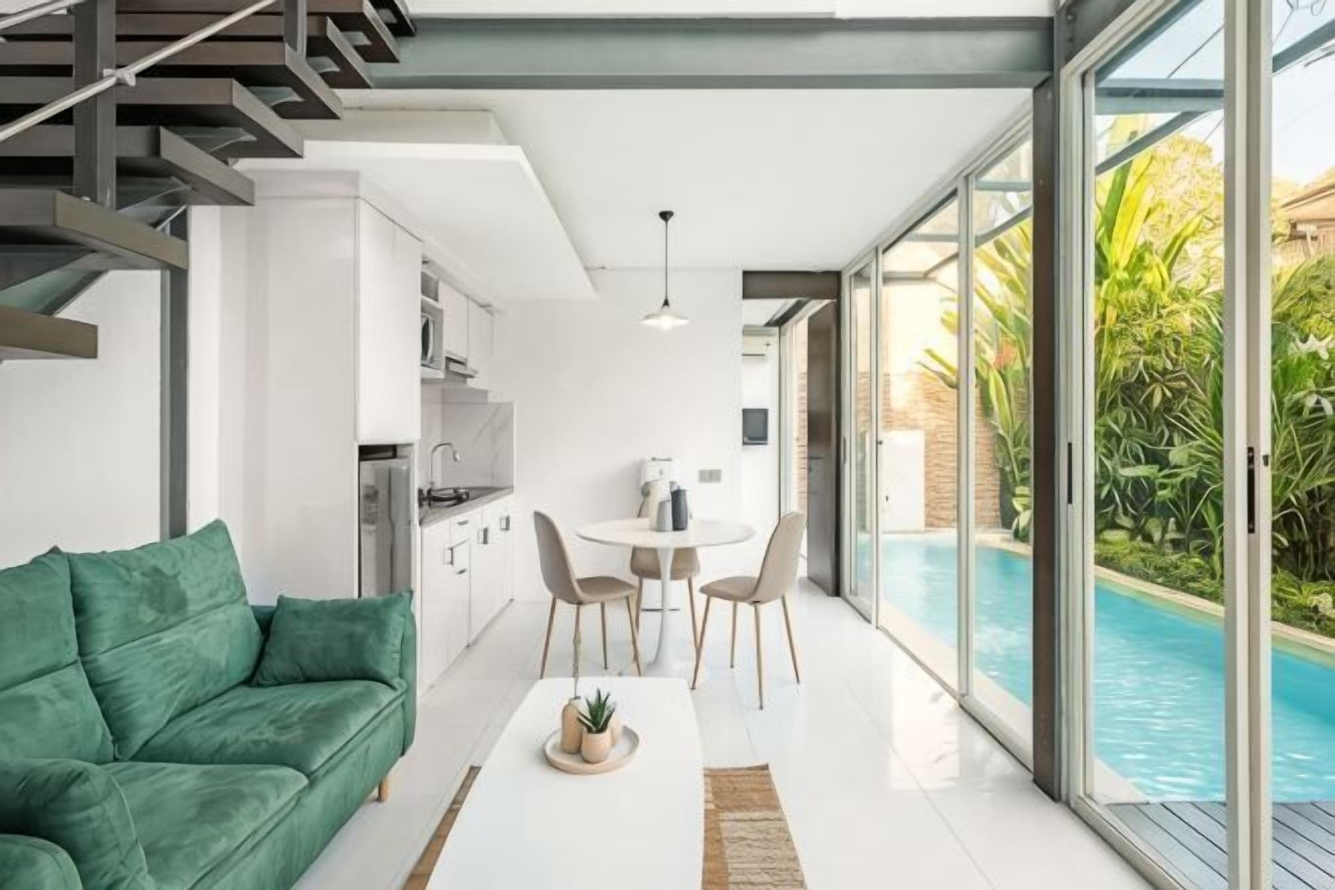 A modern living room with a green sofa, a dining table with chairs, a kitchen area, and a poolside view.