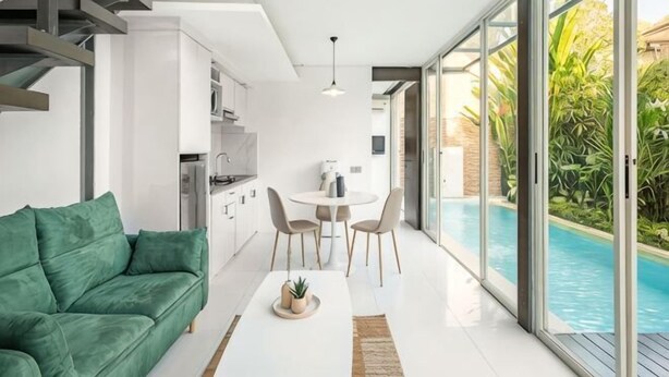A modern living room with a green sofa, a dining table with chairs, a kitchen area, and a poolside view. - Villa De Luca By Aishwarya Bali International (Bali)