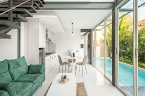 Een moderne woonkamer met een groene bank, een eettafel met stoelen, een keuken en uitzicht op het zwembad.