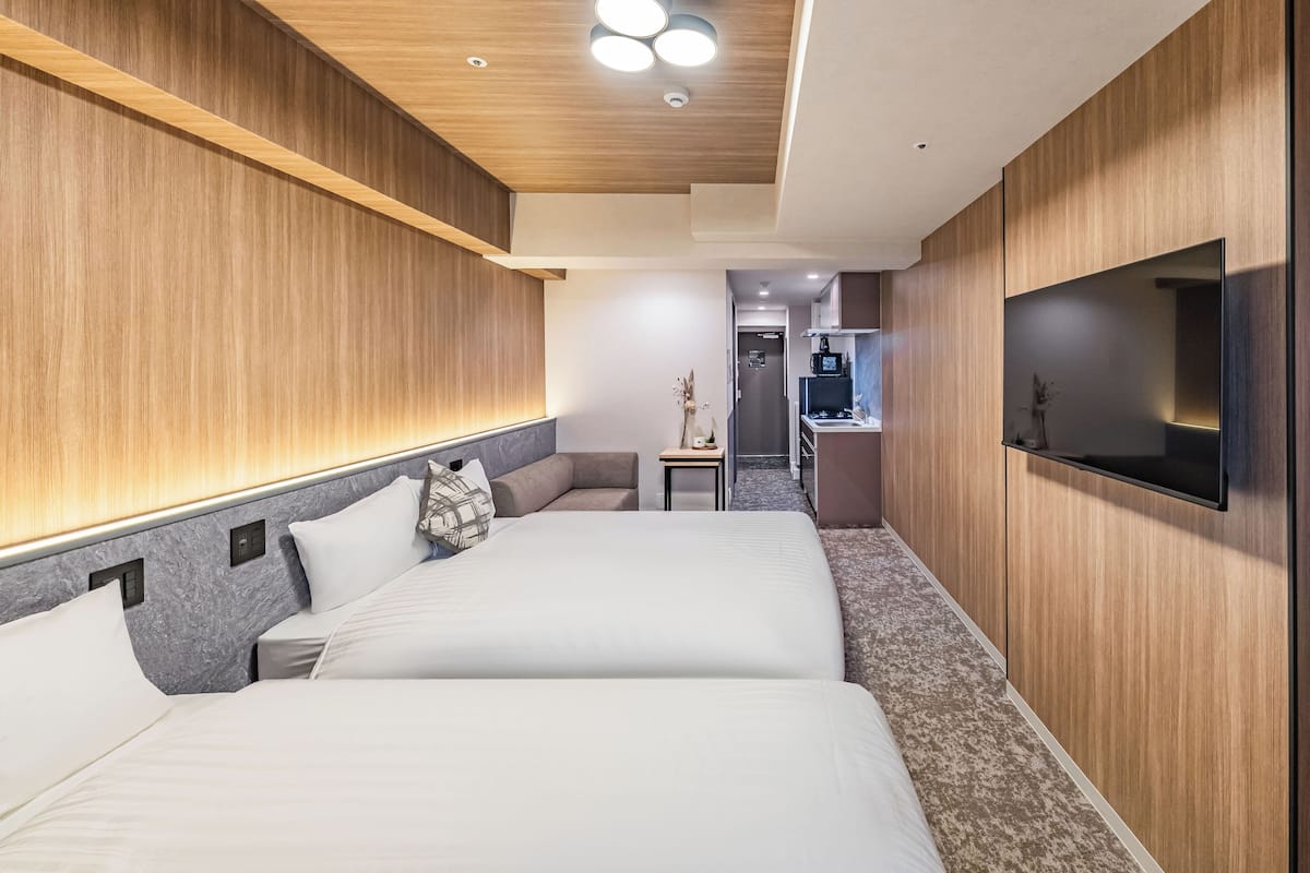 Una habitación de hotel moderna con dos camas, un pequeño rincón de descanso, un televisor de pantalla plana y una kitchenette al fondo.
