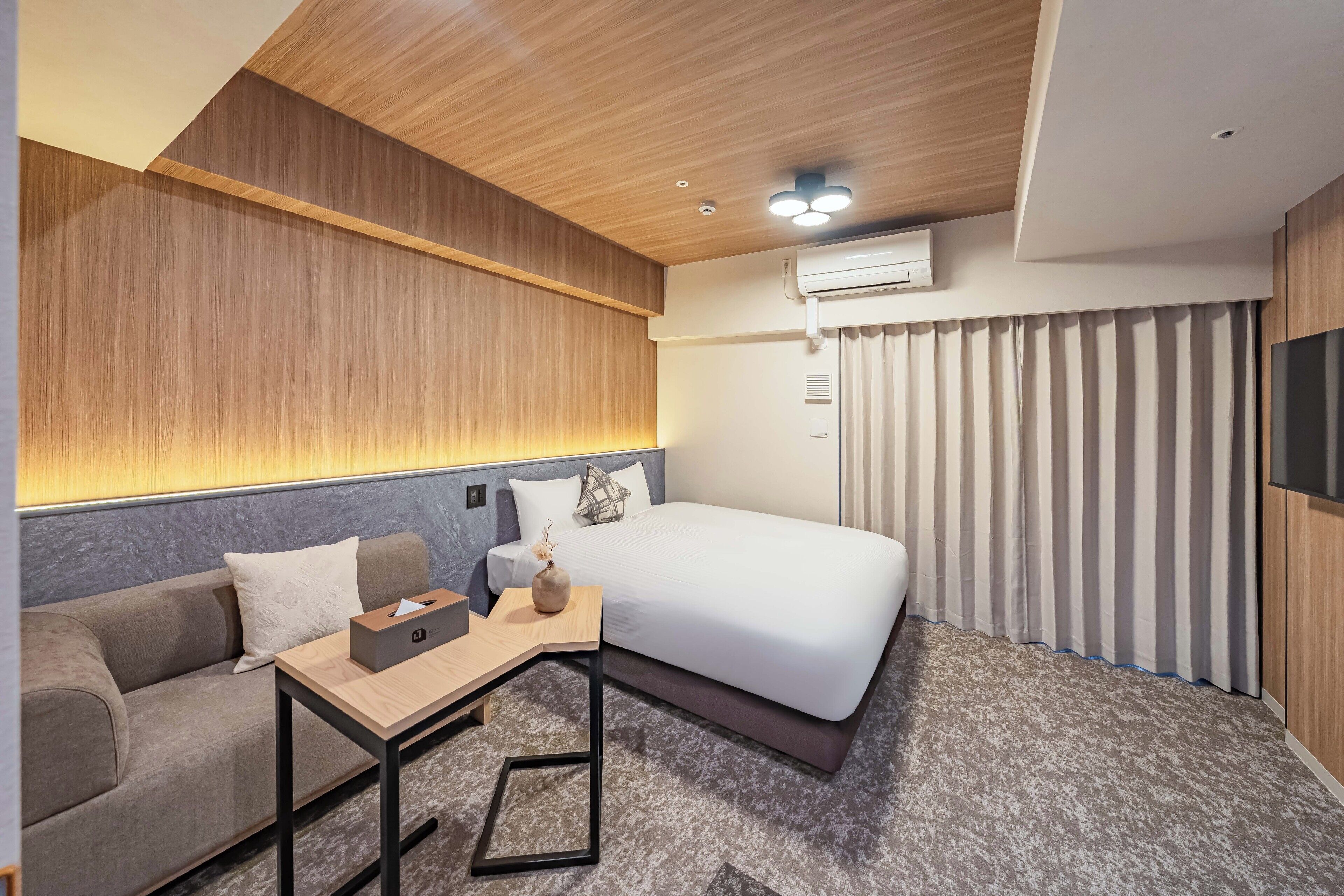 A modern hotel room with a wooden ceiling, a bed with white bedding, a grey sofa, a small table with a vase, and a TV on the wall.