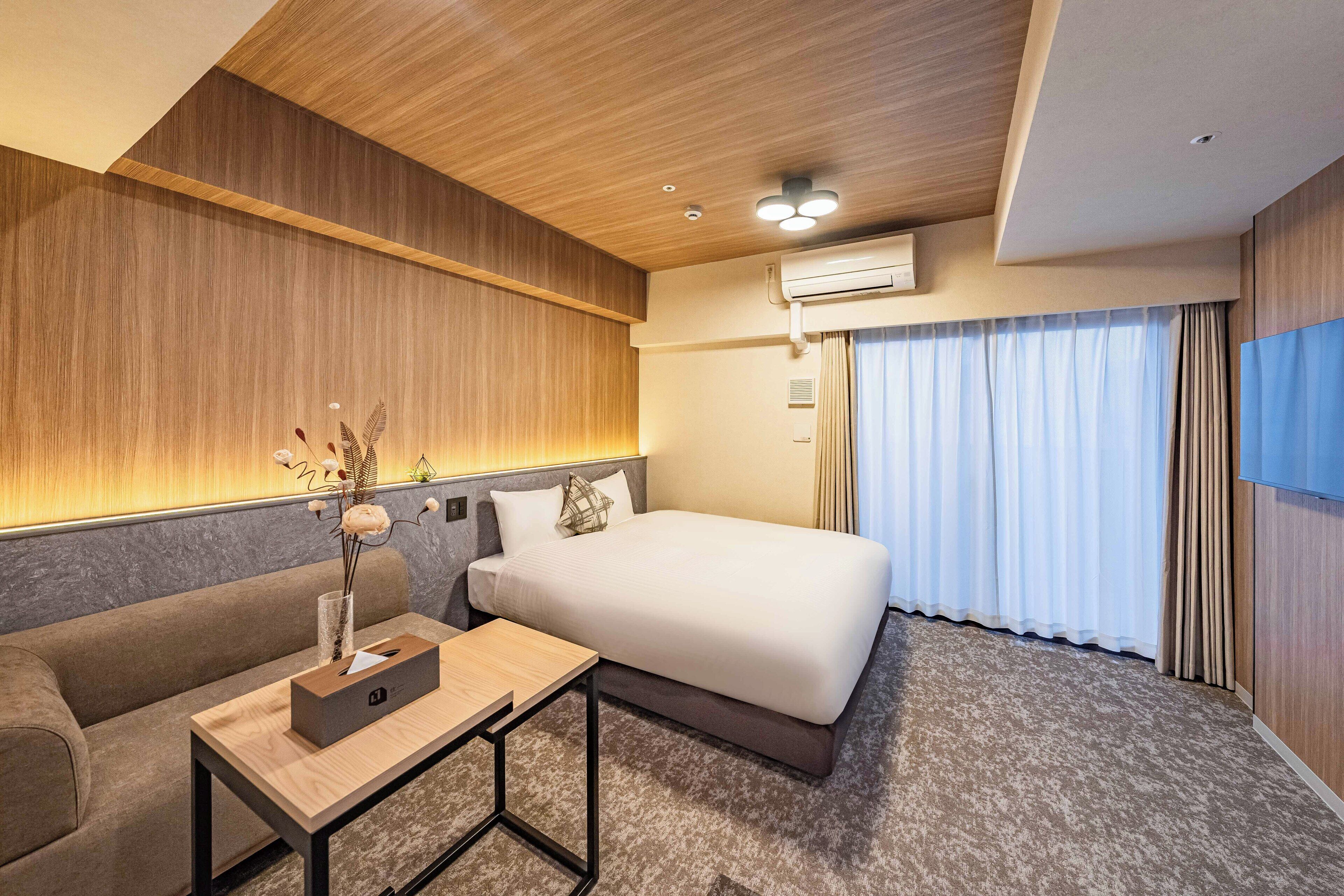A modern hotel room with a wooden ceiling, a bed with a white mattress, a grey sofa, a small wooden table with a box and a vase, and a window with curtains.