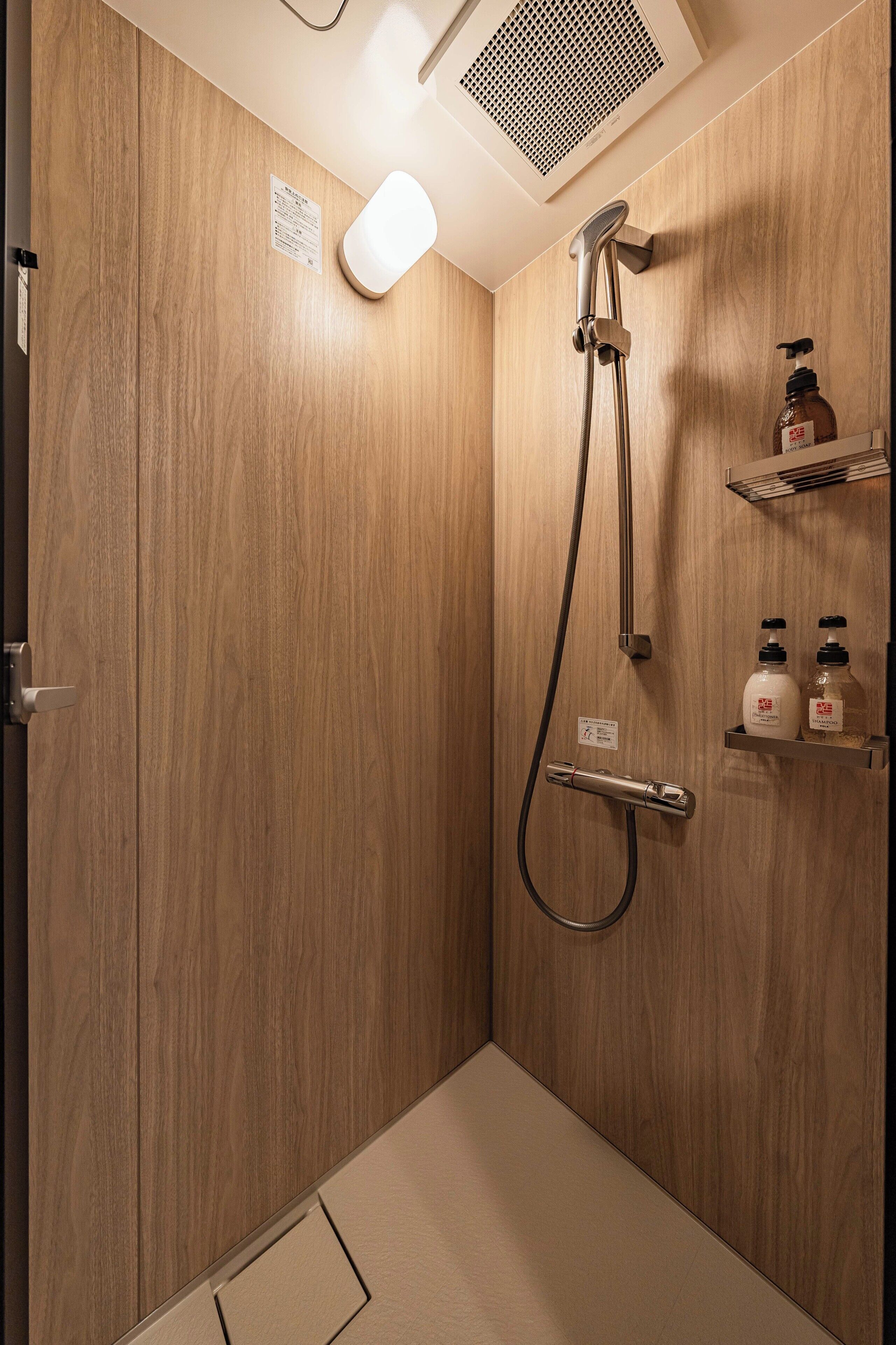 Eine Dusche mit Holzverkleidung, einer Handbrause, einem Wandarmaturen-Set und zwei Seifenspendern auf einem Regal.