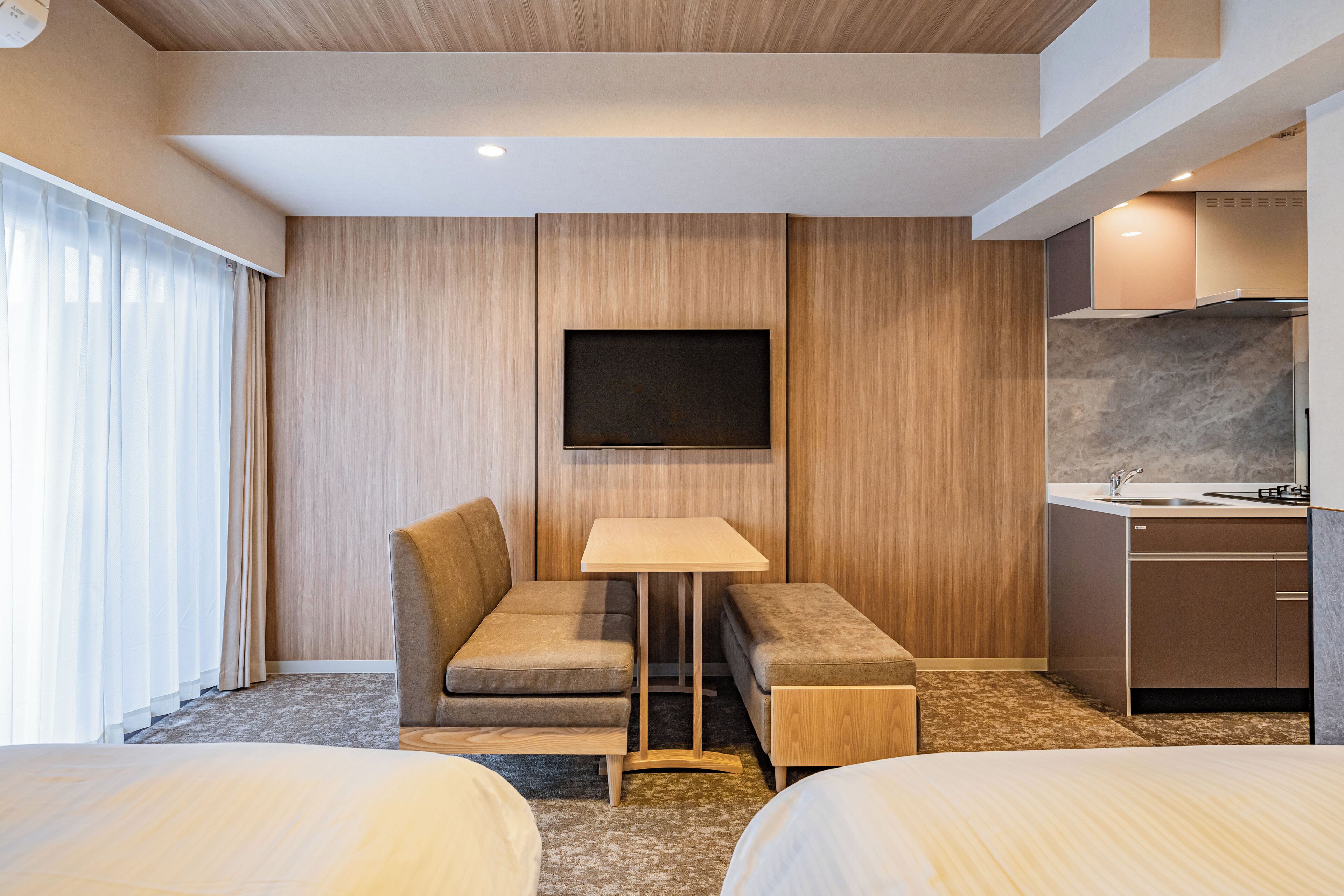 A hotel room with two beds, a small sofa, a desk, a flat-screen TV, a kitchen area with a sink and stove, and wooden panel walls.