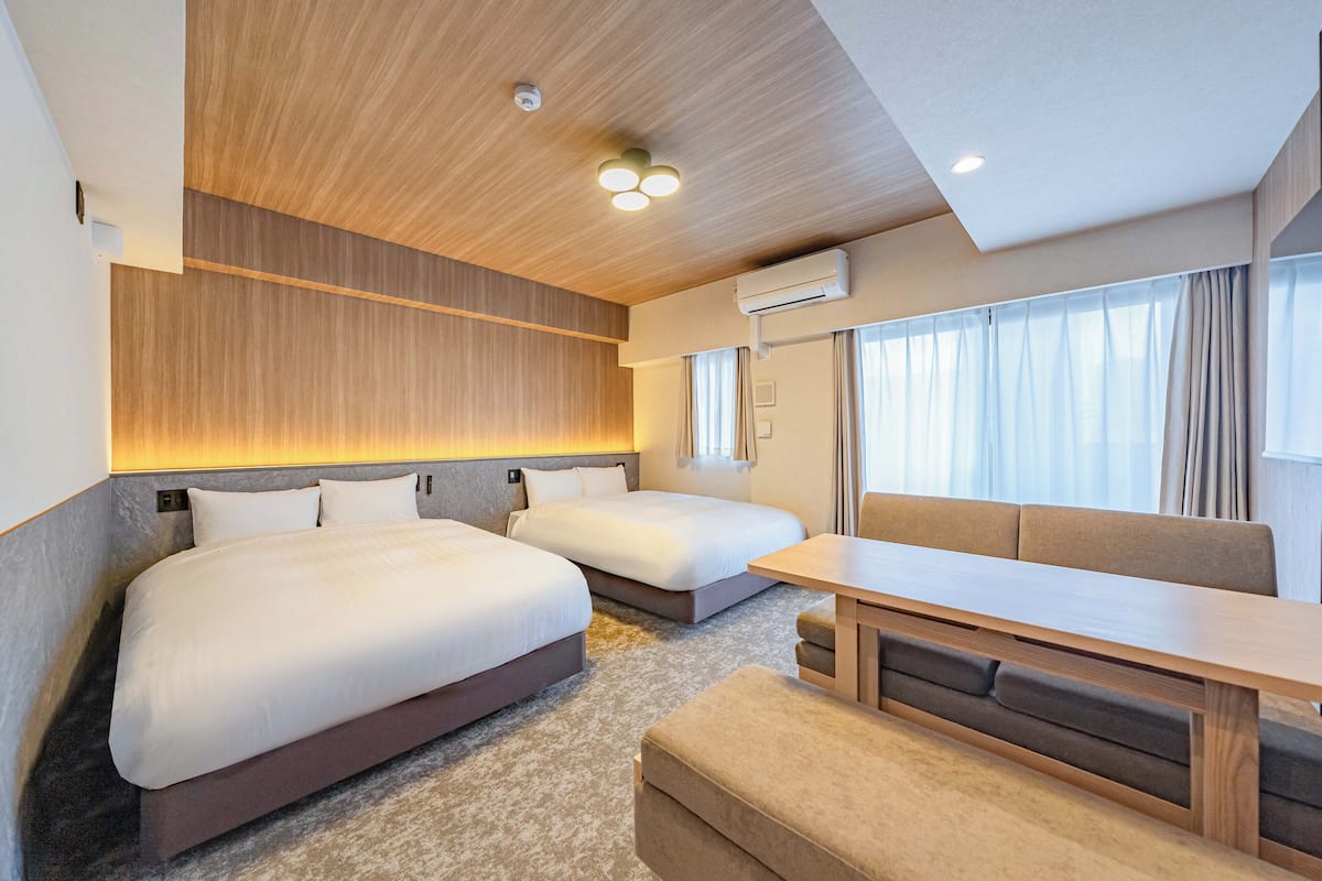 Una habitación de hotel moderna con dos camas, un banco, una mesa y un amplio ventanal con cortinas.