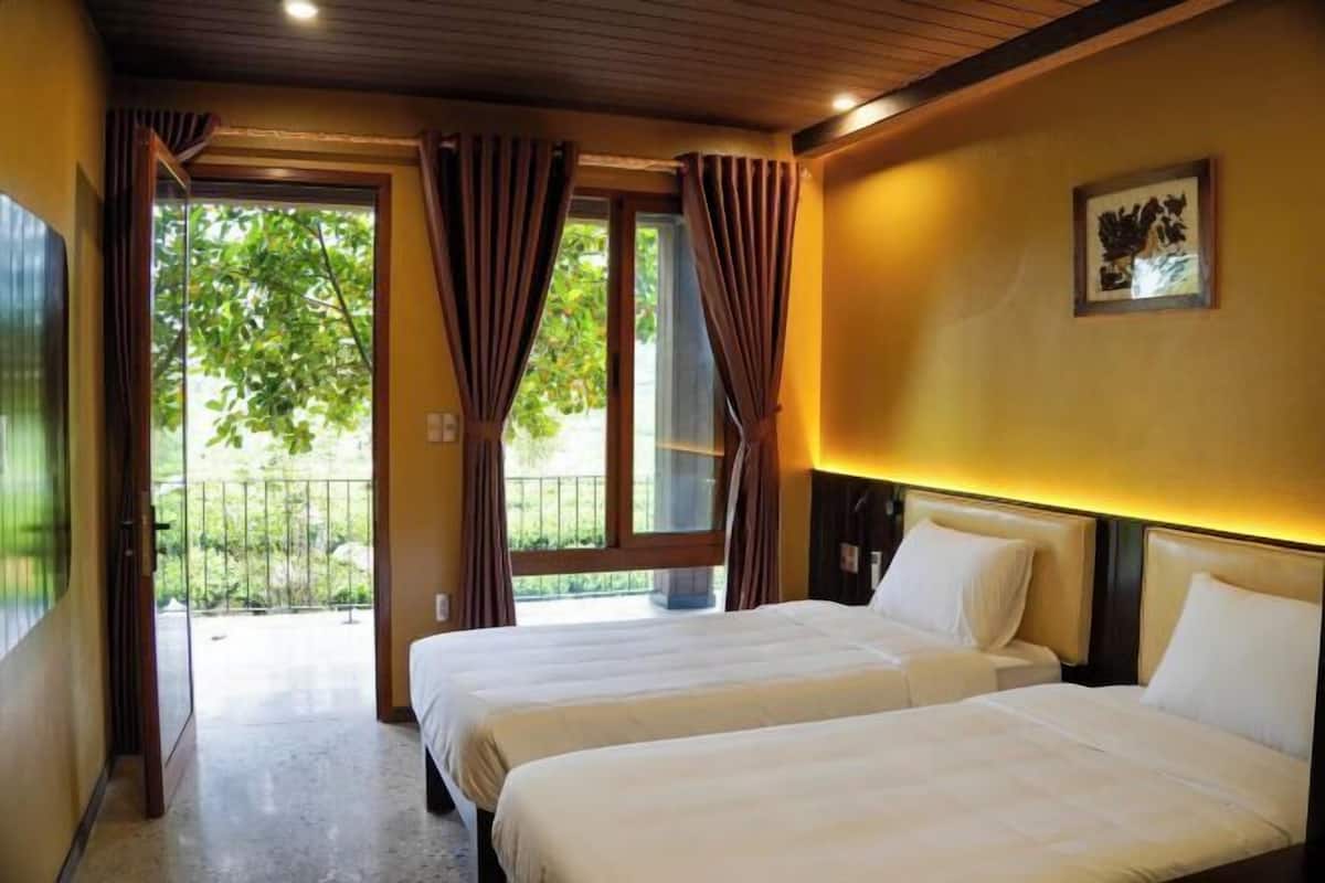 Ein Hotelzimmer mit zwei Betten, einem Balkon mit Vorhängen und Blick auf Grünfläche.