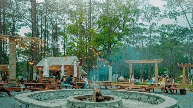 Un área de comedor al aire libre con mesas y bancos de madera, rodeada de muros de piedra y un brasero central.