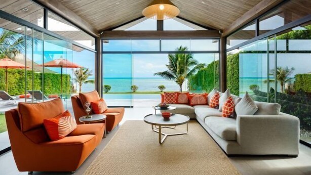 A modern living room with a large glass wall offering a view of the ocean and palm trees. - Baan Talay BeachFront Pool Villa A (Koh Samui)