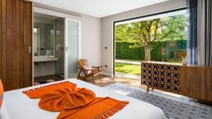 A modern bedroom with a large bed, a wooden wardrobe, a glass shower enclosure, and a view of a garden through a sliding glass door. - Baan Talay BeachFront Pool Villa A (Koh Samui)
