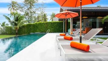 Zona junto a la piscina con sillas de descanso y una sombrilla, rodeada de setos cuidados y palmeras.