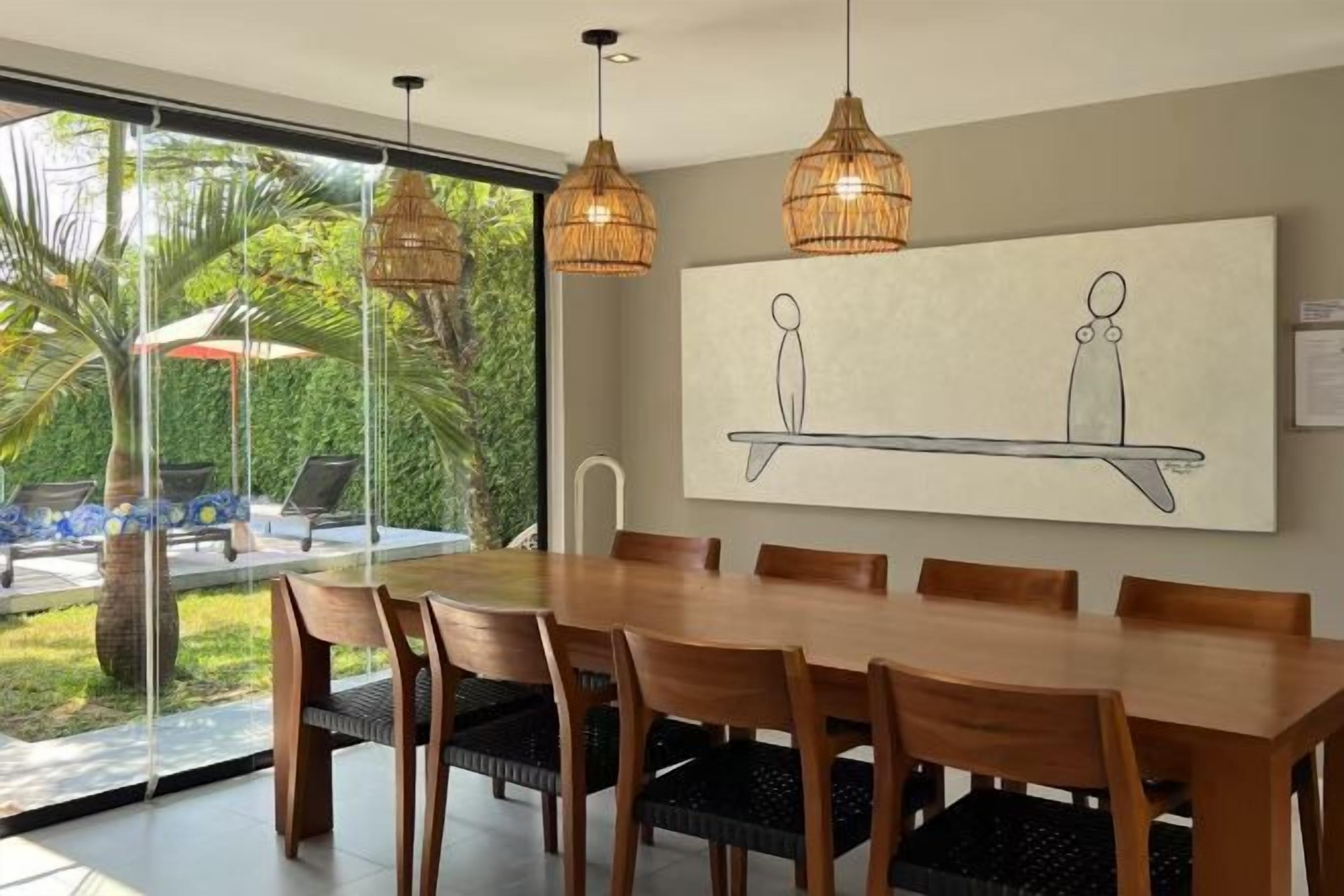 Un comedor con una gran mesa de madera y sillas, dos lámparas colgantes de mimbre y un cuadro abstracto de gran tamaño en la pared.