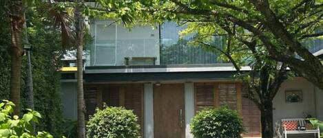A modern house with a glass balcony, a wooden door, and a small garden with two bushes.