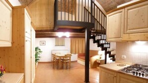 A kitchen with wooden cabinets, a gas stove, a dining area with a table and chairs, and a staircase with a black railing. - Apartment 39 for Families or Couples (Castel del Piano)
