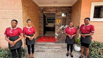 Four individuals in red uniforms standing in front of a building entrance with a red carpet and a sign indicating 'House 1'.