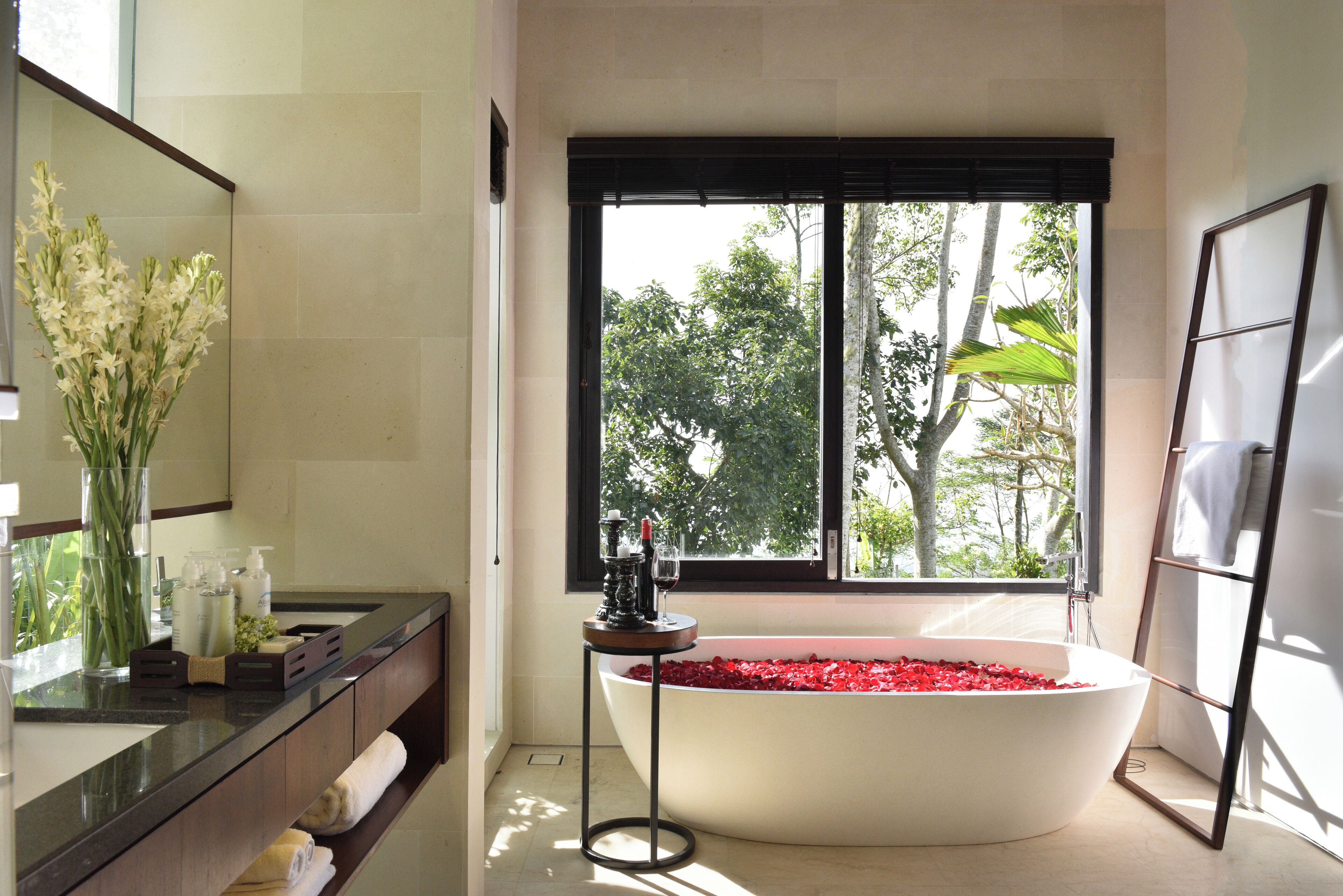A modern bathroom with a freestanding bathtub filled with rose petals, a vanity with a mirror, and a window with a view of trees.