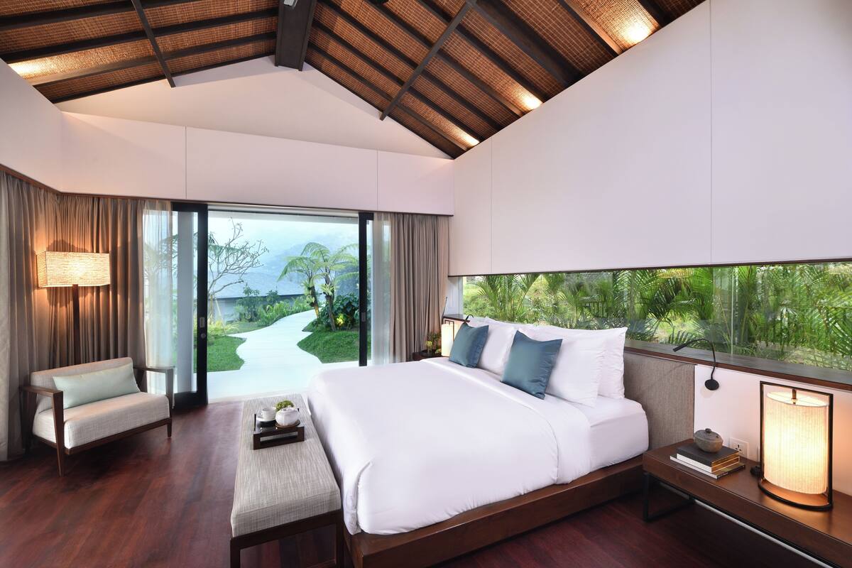 A modern bedroom with a large bed, a sofa, a bedside table with books, and a view of a tropical landscape.