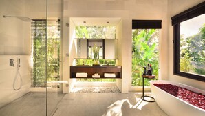 A modern bathroom with a glass shower enclosure, a wooden vanity with a mirror, and a large window offering a view of greenery. - ALTA VISTA BALI MOUNTAIN VILLAS (Wanagiri)