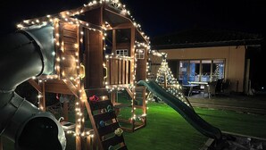 Una casita de juegos de madera con un tobogán, rodeada de luces de cuerda, sobre un césped.