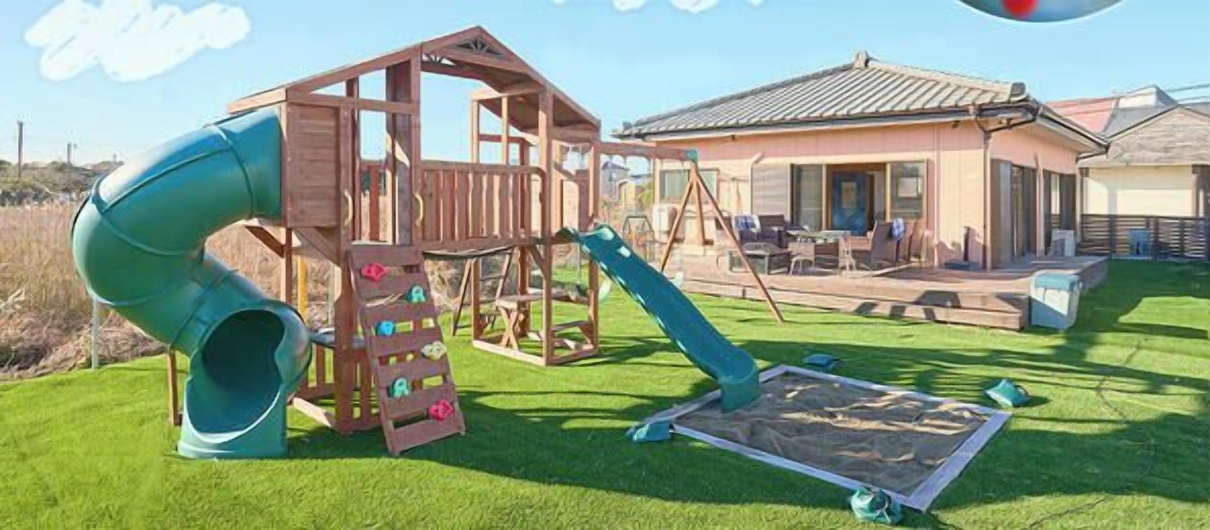 A wooden play structure with a slide and climbing steps, a covered seating area, and a house in the background.