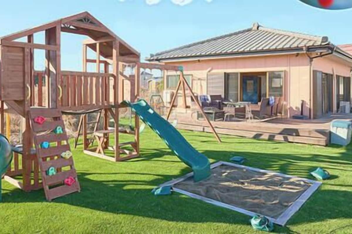 Una estructura de juego de madera con un tobogán y escalones de acceso, un área de descanso cubierta y una casa al fondo.