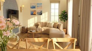 A modern living room with a sectional sofa, a coffee table, and a TV. There are wall-mounted lights and a large window with curtains. - Novaworld Phan Thiet - Alala Stay (Phan Thiet)