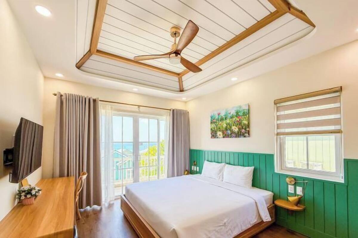 Une chambre d’hôtel avec un lit, un bureau, un ventilateur de plafond, un tableau et une fenêtre.