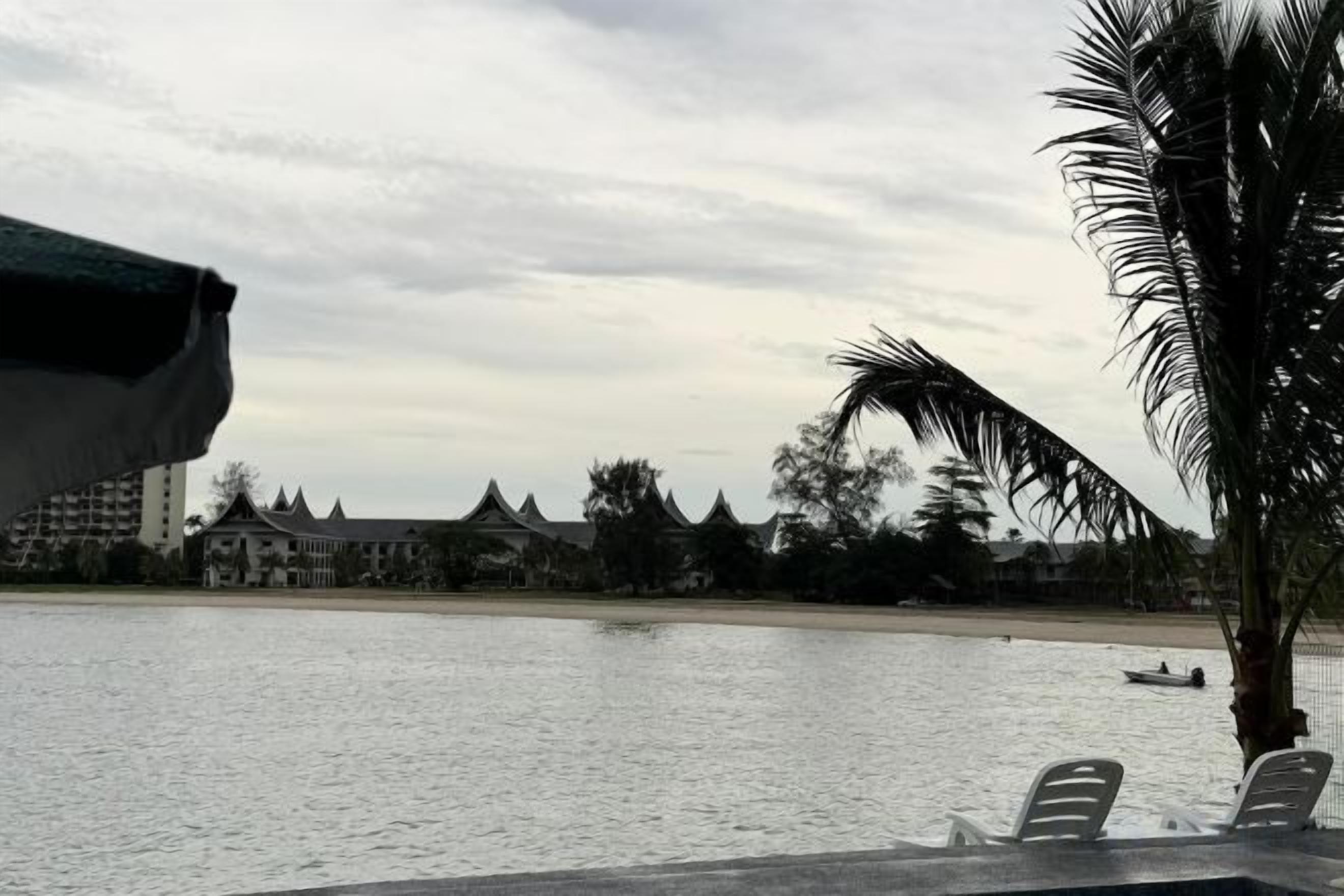 A view of a beach with a palm tree, a boat, and a building in the background.