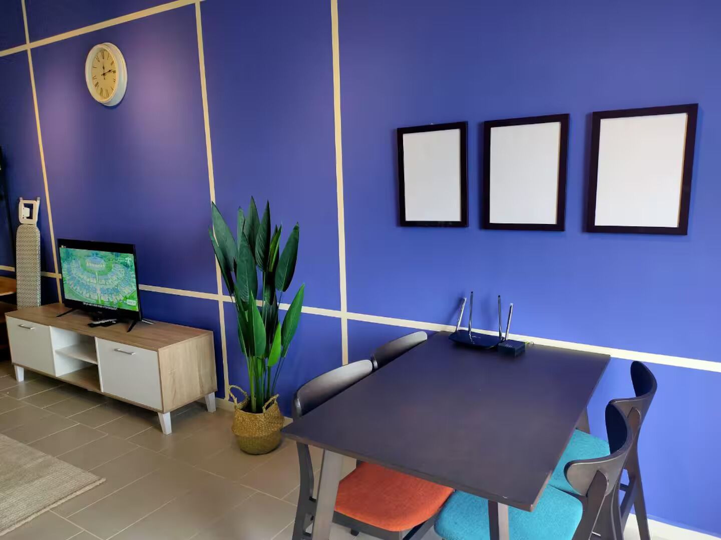 A room with a blue wall, a clock, a television, a plant, a table with chairs, and three empty picture frames.