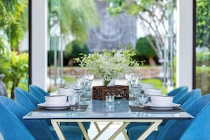 A dining table set with glassware and plates, surrounded by blue upholstered chairs, in a room with a view of greenery. - Turquoise Villa 2 GYM and KIDS ROOM (Koh Samui)