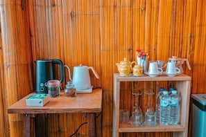 A bamboo wall, a wooden table with a kettle, teapot, cups, and a shelf with bottles and glasses.