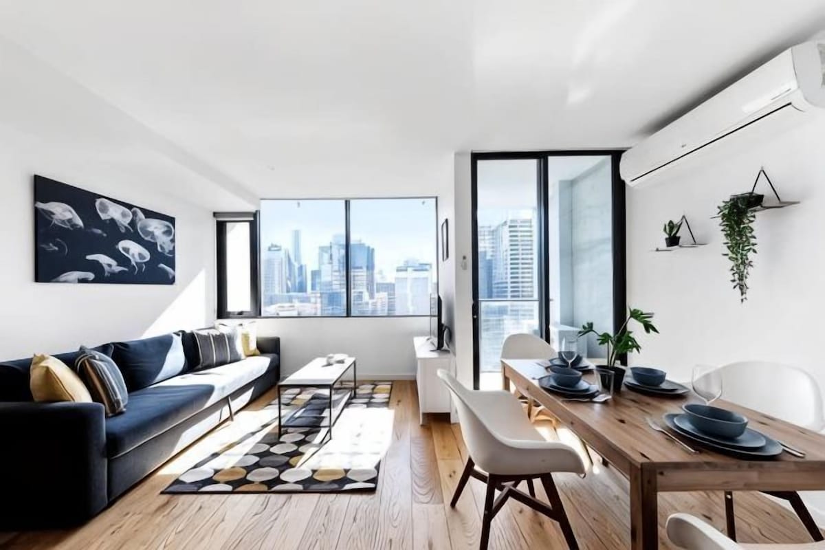 A modern living room with a sofa, dining table, chairs, and a city view through large windows.