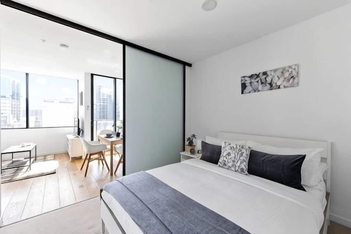 Une chambre moderne avec un grand lit, un bureau, une chaise et une vue sur la ville grâce à de grandes fenêtres.