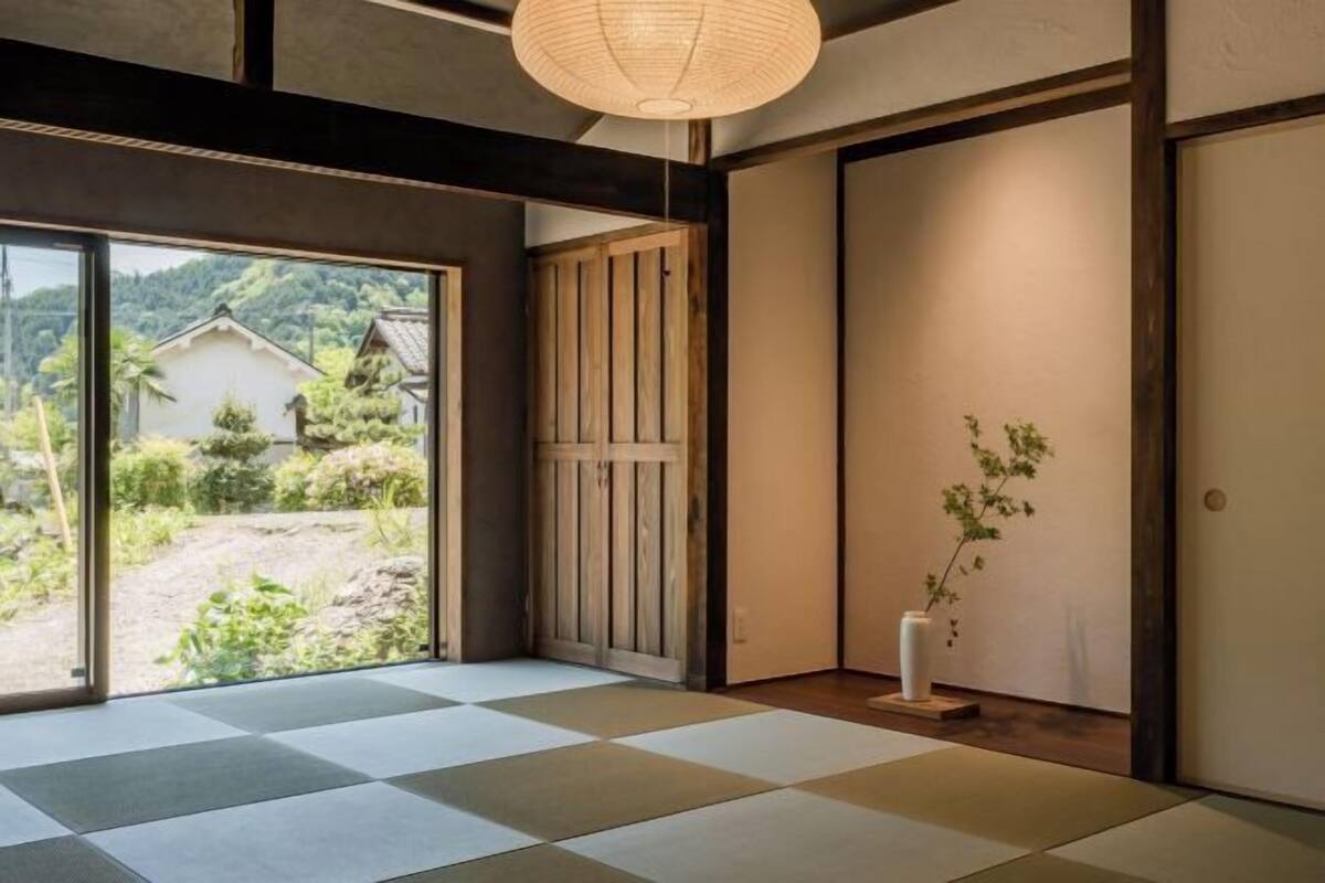 Una habitación con suelo de tatami, una puerta corredera, una pequeña planta de interior y vistas a un jardín y edificios.