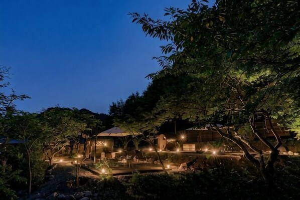 A well-lit outdoor area with a covered seating area, surrounded by trees and a building with a sloped roof.