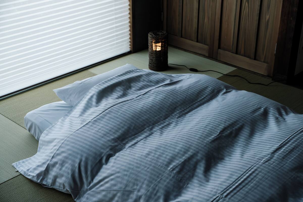 Una habitación con suelo de tatami, una cama con manta a rayas, una mesita con una vela y una ventana con persianas.